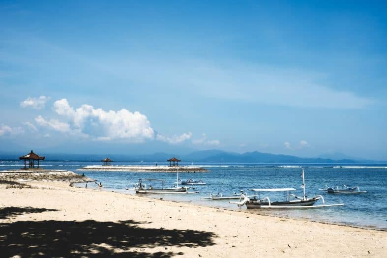 white sand beach in sanur with traditional boats and the coastline in the distance